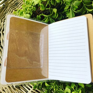 May include: A small, brown notebook with a clear plastic pocket on the front cover. The notebook has lined pages and is open to reveal the blank pages.