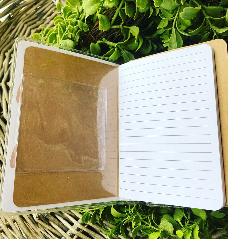 May include: A small, brown notebook with a clear plastic pocket on the front cover. The notebook has lined pages and is open to reveal the blank pages.