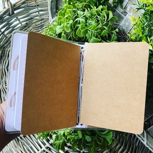May include: A brown notebook with a clear plastic cover, open to reveal blank pages. The notebook is lying on a wicker surface.