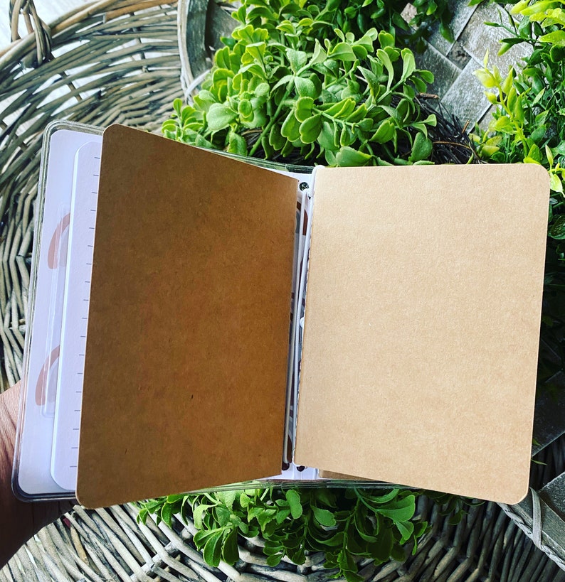 May include: A brown notebook with a clear plastic cover, open to reveal blank pages. The notebook is lying on a wicker surface.