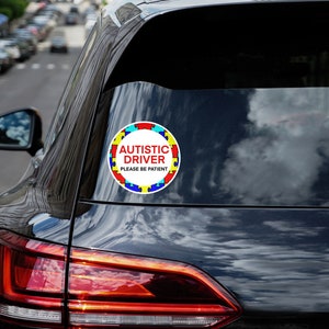 K&ouml;nnte beinhalten: Ein runder wei&szlig;er Aufkleber mit einem Rand aus blauen, gelben und roten Puzzle-Teilen. Der Aufkleber lautet "Autistic Driver Please Be Patient" in roter Schrift.