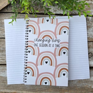 May include: A white spiral bound notebook with a pink, brown, and white rainbow pattern. The notebook has the text "changing lives one session at a time" on the front cover.