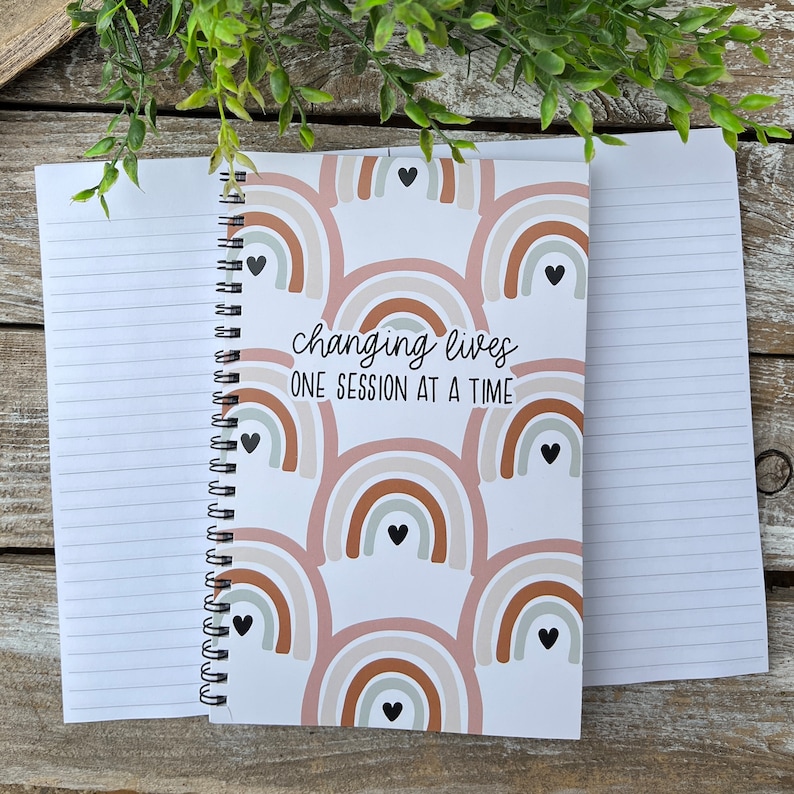 May include: A white spiral bound notebook with a pink, brown, and white rainbow pattern. The notebook has the text "changing lives one session at a time" on the front cover.