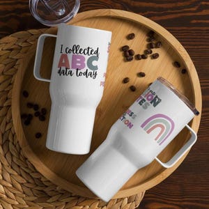 May include: Two white travel mugs with different designs. The first mug has the text "I collected ABC data today" in black lettering with the letters ABC in pink, blue, and green. The second mug has a rainbow design with the text "It's all about the education" in purple lettering.
