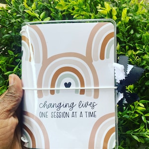 May include: A white planner with a clear plastic cover. The cover features a rainbow pattern in shades of brown, beige, and green. The planner has a white elastic band and the text "changing lives one session at a time" is printed on the front.