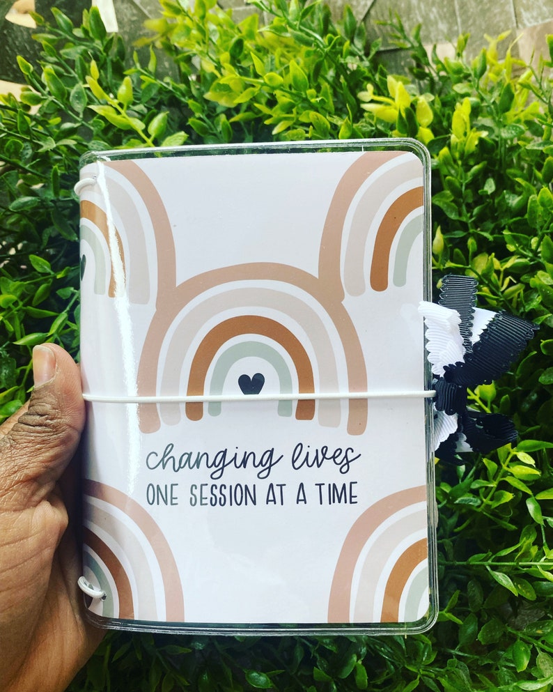 May include: A white planner with a clear plastic cover. The cover features a rainbow pattern in shades of brown, beige, and green. The planner has a white elastic band and the text "changing lives one session at a time" is printed on the front.