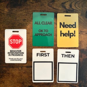 Puede incluir: Un conjunto de letreros rectangulares con varios mensajes. Un letrero dice "STOP" con "BEHAVIOR INTERVENTION IN PROGRESS" debajo. Otros letreros dicen "ALL CLEAR", "Need help!", "FIRST" y "THEN". Cada letrero tiene un agujero en la parte superior.