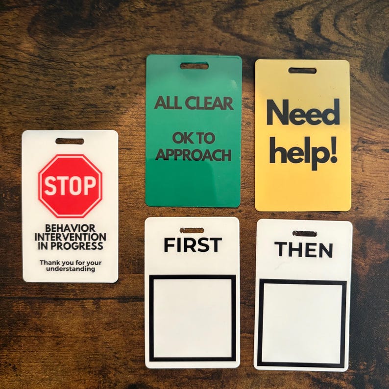 Puede incluir: Un conjunto de letreros rectangulares con varios mensajes. Un letrero dice "STOP" con "BEHAVIOR INTERVENTION IN PROGRESS" debajo. Otros letreros dicen "ALL CLEAR", "Need help!", "FIRST" y "THEN". Cada letrero tiene un agujero en la parte superior.