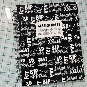 May include: Black and white spiral bound notebook with the words "Session Notes: Changing Lives, One Session at a Time" on a sticker. The cover is covered in a repeating pattern of the words "ABA, BIP, FCT, SD, SEAT, behavior, analysis, data, changing lives, applied".