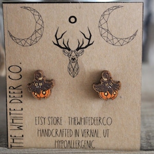 May include: Wooden Halloween-themed stud earrings. Each earring features a brown bat perched atop an orange pumpkin. The earrings are displayed on a brown card with the text "THE WHITE DEER CO." and "HANDCRAFTED IN VERNAL, UT HYPOALLERGENIC."