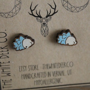 May include: A pair of wooden earrings featuring a blue and white cartoon hedgehog design. The earrings are displayed on a brown card with the text "The White Deer Co." and "Etsy Store: thewhitedeerco Handmade in Vernal, UT Hypoallergenic".