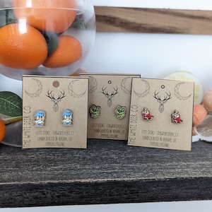 Könnte beinhalten: Drei Sätze kleiner, hölzerner Ohrringe mit verschiedenen Designs. Das erste Set zeigt zwei blaue Vögel, das zweite Set zeigt zwei grüne Dinosaurier und das dritte Set zeigt zwei rote Drachen. Jedes Set wird auf einer kleinen, braunen Karte mit dem Text "The White Deer Co." und "Etsy Store: TheWhiteDeerCo Handcrafted in Vernal UT Hypoallergenic" angezeigt.