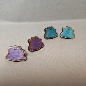 May include: Four wooden stud earrings in a whimsical design. Two earrings are teal, and two are purple. Each earring has a gold-colored border and a small, curved indentation at the top. The earrings are displayed on a light gray surface.