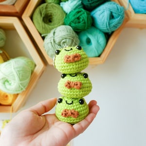 May include: Three crocheted green frog plushies stacked on top of each other. Each frog has black eyes, a brown bow tie, and a smiling face.