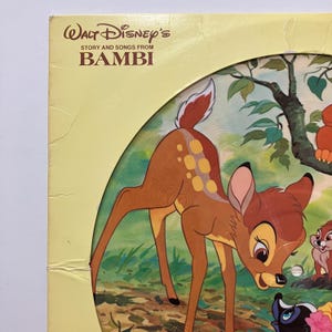May include: A vintage Walt Disney's "Bambi" story and songs record album cover. The cover features a brown deer with yellow spots, a pink-eared deer, and other forest creatures. The album title is in brown text on a yellow background.