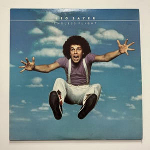 May include: Album cover for "Endless Flight" by Leo Sayer. The image features a man mid-air, arms outstretched, against a blue sky with clouds. He wears a purple shirt, white pants with suspenders, and black shoes. The album title and artist name are at the top.