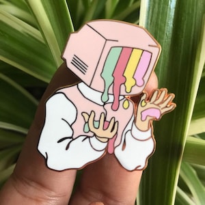 Op de afbeelding: Een roze emaille pin van een persoon met een televisiescherm als hoofd. Het scherm smelt met kleurrijke druppels groen, geel, roze en oranje. De persoon draagt een wit shirt en heeft een hand uitgestoken.