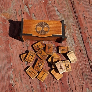 Puede incluir: Caja de madera con un diseño de árbol de la vida en la tapa, abierta para revelar un conjunto de runas de madera con símbolos nórdicos.