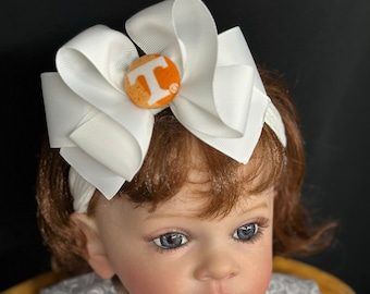 Peut inclure: Une poupée bébé portant un bandeau blanc avec un grand nœud blanc et un bouton orange avec un T blanc. La poupée porte une robe à motifs gris et blancs avec une bordure de volants blancs et un motif à carreaux orange et blancs en dessous.