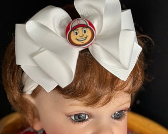May include: A white bow headband with a red and white Ohio State Buckeyes button in the center. The headband is on a baby doll with brown hair and blue eyes.