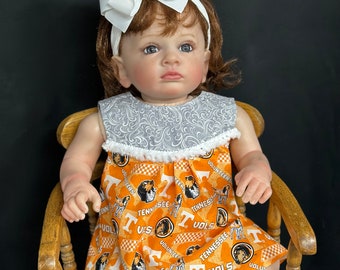 May include: A baby doll wearing a white headband with an orange 'T' on a bow, and an orange and white dress with a checkered pattern and the words 'Tennessee Vols' repeated throughout.