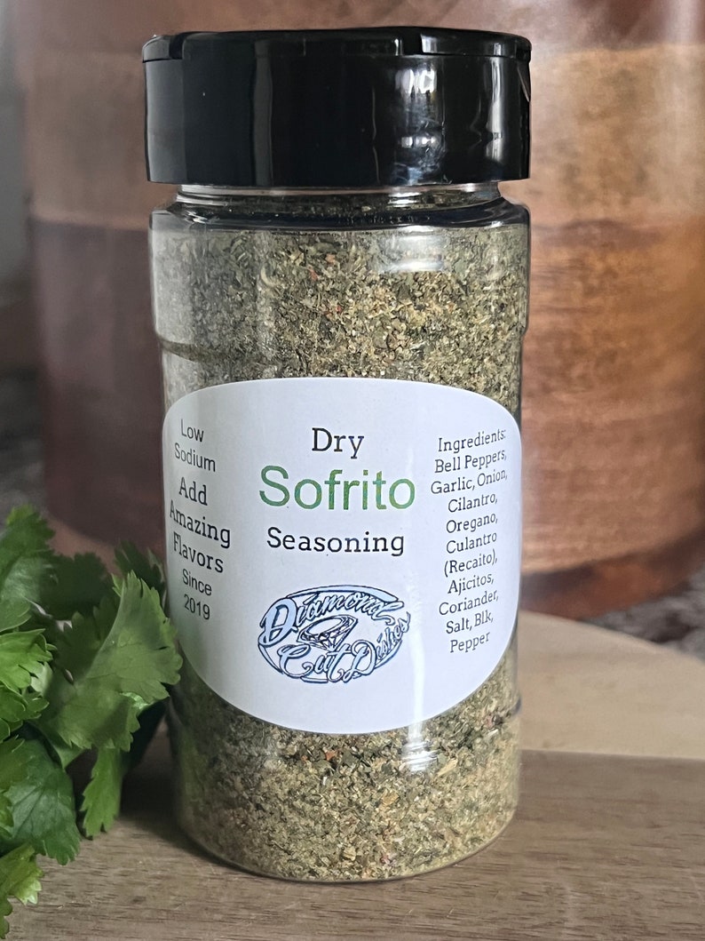 May include: A clear glass jar with a black lid filled with dried herbs and spices. The label reads "Dry Sofrito Seasoning" and lists the ingredients: Bell Peppers, Garlic, Onion, Cilantro, Oregano, Culantro (Recaito), Ajicitos, Coriander, Salt, Black Pepper. The label also says "Low Sodium, Add Amazing Flavors, Since 2019".