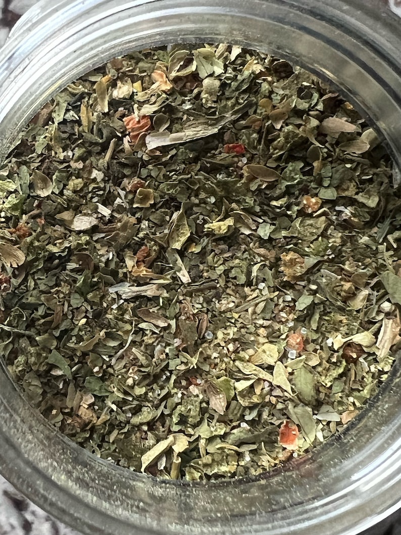 May include: A close-up view of a glass jar filled with a blend of dried herbs and spices. The mixture is a mix of green, brown, and red pieces, with a few small white specks.