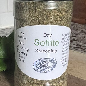 May include: A clear glass jar with a black lid filled with dried herbs and spices. The label reads "Dry Sofrito Seasoning" and includes the text "Low Sodium, Add Amazing Flavors, Since 2019".