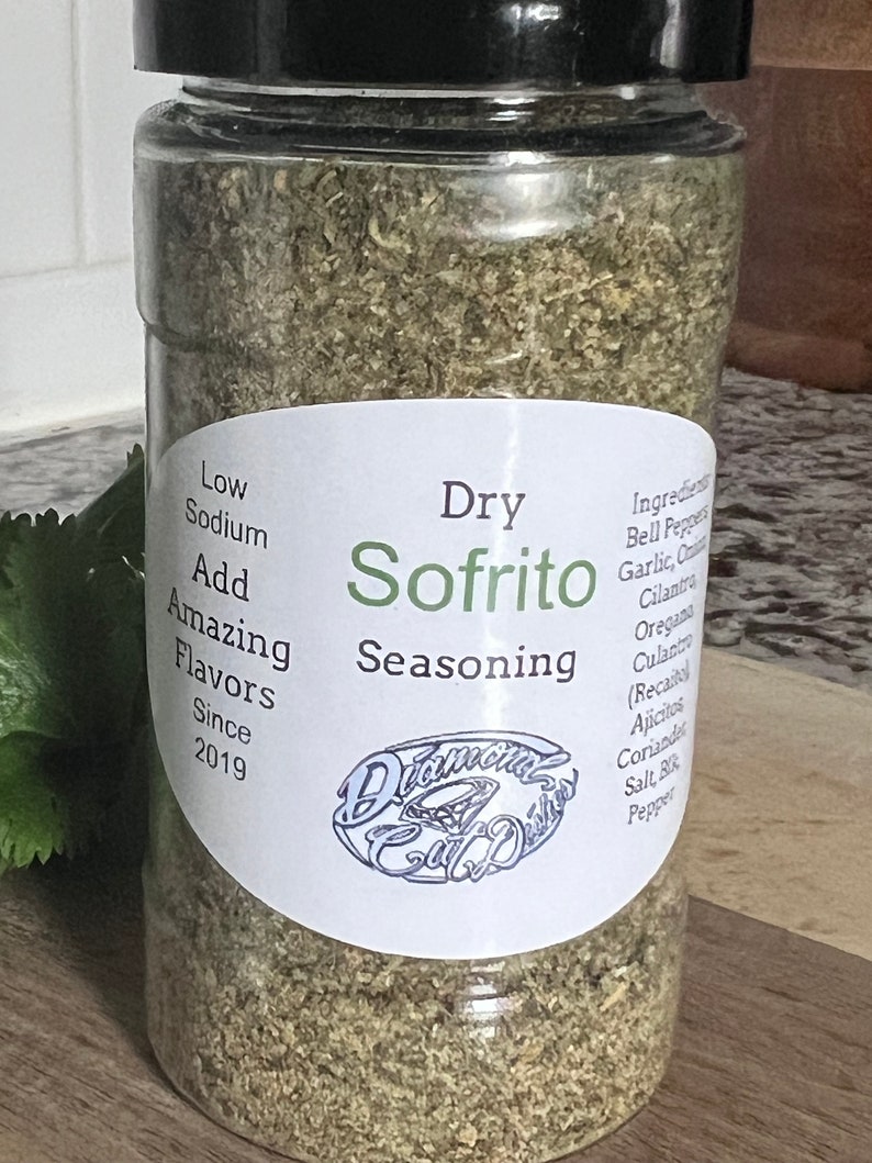 May include: A clear glass jar with a black lid filled with dried herbs and spices. The label reads "Dry Sofrito Seasoning" and includes the text "Low Sodium, Add Amazing Flavors, Since 2019".