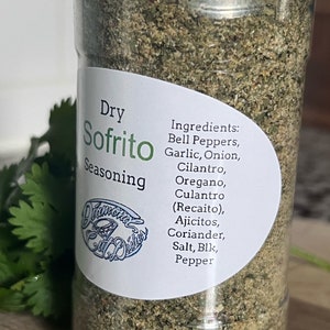 May include: A glass jar filled with a green, dry sofrito seasoning blend. The label reads "Dry Sofrito Seasoning" and lists the ingredients: Bell Peppers, Garlic, Onion, Cilantro, Oregano, Culantro (Recaito), Ajicitos, Coriander, Salt, Blk, Pepper.