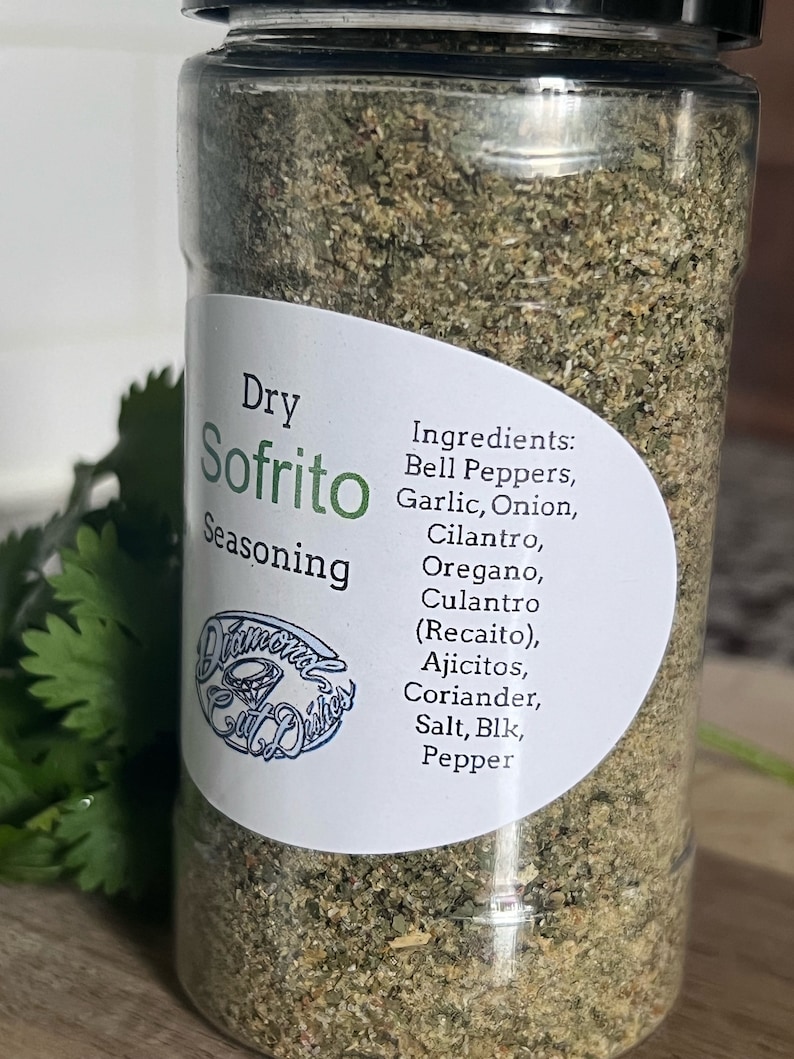 May include: A glass jar filled with a green, dry sofrito seasoning blend. The label reads "Dry Sofrito Seasoning" and lists the ingredients: Bell Peppers, Garlic, Onion, Cilantro, Oregano, Culantro (Recaito), Ajicitos, Coriander, Salt, Blk, Pepper.