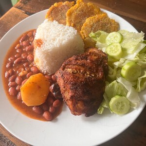 May include: A white plate with a meal of roasted chicken, white rice, red beans, fried plantains, and a side salad of lettuce and cucumber.