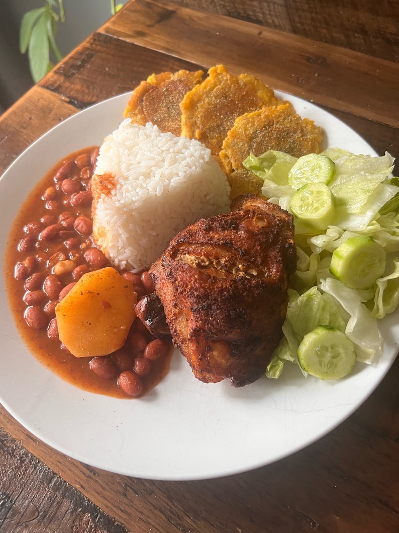 May include: A white plate with a meal of roasted chicken, white rice, red beans, fried plantains, and a side salad of lettuce and cucumber.