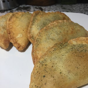 May include: Five golden brown empanadas, some sprinkled with herbs, are arranged on a white plate.
