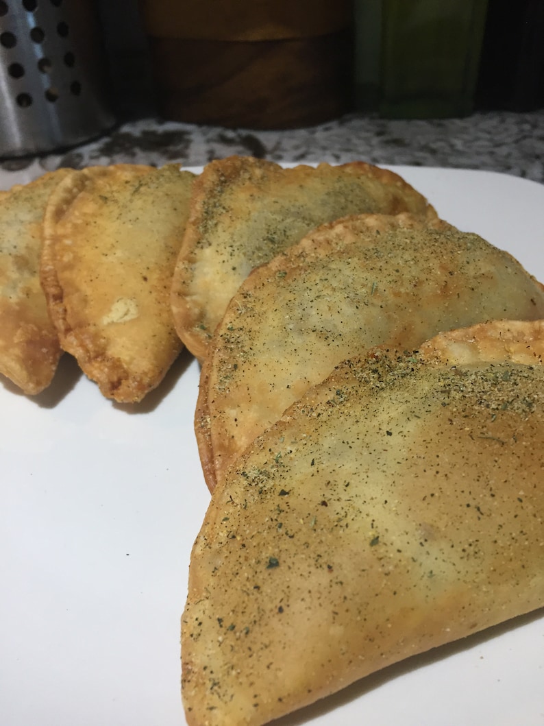 May include: Five golden brown empanadas, some sprinkled with herbs, are arranged on a white plate.