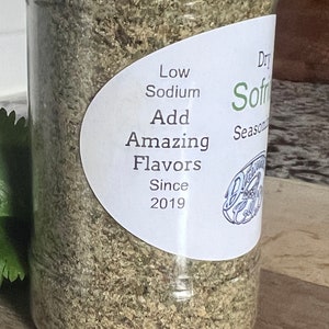 May include: A clear plastic jar filled with a light green and brown seasoning blend. The label reads "Dry Sofrito Seasoning", "Low Sodium", "Add Amazing Flavors", and "Since 2019".