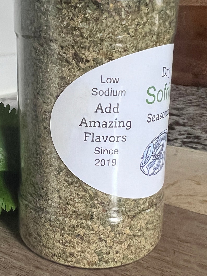 May include: A clear plastic jar filled with a light green and brown seasoning blend. The label reads "Dry Sofrito Seasoning", "Low Sodium", "Add Amazing Flavors", and "Since 2019".
