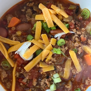 May include: A bowl of chili topped with shredded cheese and chopped green onions. The chili is a rich, red color and appears to be made with ground beef, beans, and vegetables.