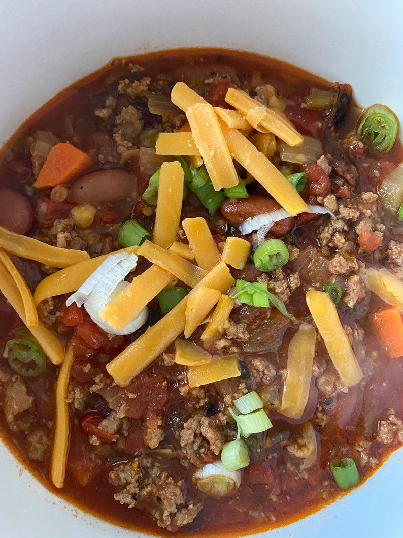 May include: A bowl of chili topped with shredded cheese and chopped green onions. The chili is a rich, red color and appears to be made with ground beef, beans, and vegetables.