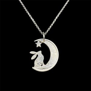 May include: A silver pendant necklace with a crescent moon design, a star, and a rabbit silhouette. The pendant hangs from a delicate silver chain. The background is black.