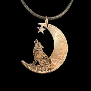 Può includere: Una collana con pendente color rame raffigurante la silhouette di un lupo che ulula intagliata da una moneta, ambientata contro una luna crescente e una stella. L'anno 1967 è visibile sulla moneta. La collana è appesa a un cordino verde scuro.