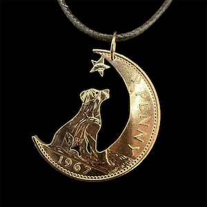 May include: A copper-colored dog pendant necklace. The pendant features a dog silhouette sitting on a crescent moon with a star above. The year 1967 is engraved on the moon. The necklace hangs on a dark cord.