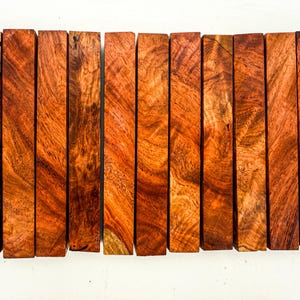 May include: A collection of eleven rectangular wood blocks with a rich, reddish-brown hue and intricate grain patterns. The blocks are arranged vertically, showcasing the natural variations in color and texture of the wood. The wood appears to be freshly cut and ready for use.