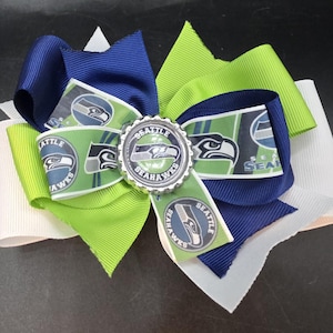 May include: A large hair bow featuring the Seattle Seahawks logo and colors. The bow is made of layered ribbons in navy blue, lime green, white, and a ribbon with the team logo. A silver embellishment is in the center.
