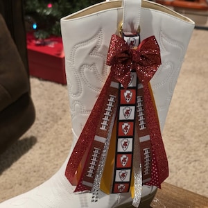 Op de afbeelding: Een witte cowboylaars met een rode strik en linten. De linten tonen voetbalpatronen en het logo van de Kansas City Chiefs. Een feestelijk accessoire voor sportfans of thema-evenementen.