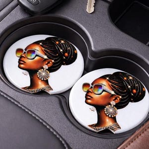 May include: Two white car coasters featuring a side profile of a woman with dark skin, wearing rainbow-tinted sunglasses and ornate jewelry. Her hair is styled in a braided updo. The coasters are placed in a car's cup holders.