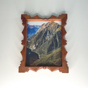 May include: A rectangular wooden picture frame with a decorative, jagged edge design. The frame contains a photograph of a steep, green mountain slope rising above a dark body of water, with rocky peaks visible in the background under a blue sky.