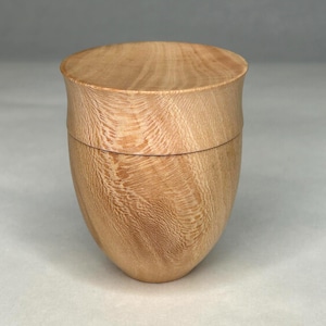 Peut inclure: Une boîte en bois faite à la main avec un couvercle. Fabriquée en bois clair, peut-être du chêne, elle présente une forme lisse et arrondie. Le couvercle s'ajuste parfaitement. Le grain du bois est visible, ajoutant à l'esthétique naturelle.