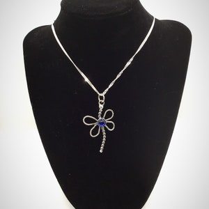 May include: A silver necklace with a dragonfly pendant featuring a blue gemstone. The necklace is displayed on a black velvet stand.
