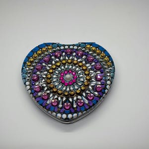 May include: Heart-shaped compact mirror with a detailed, colorful design. The compact features a blue border, a central pattern of gold and silver beads, and pink and purple gems. A central pearl adds a final touch.
